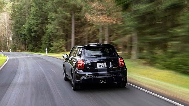 Side view of a MINI John Cooper Works 1T06 Edition vehicle driving alone on a street facing left, with bushes in the background along and the vehicle’s surroundings blurred out.