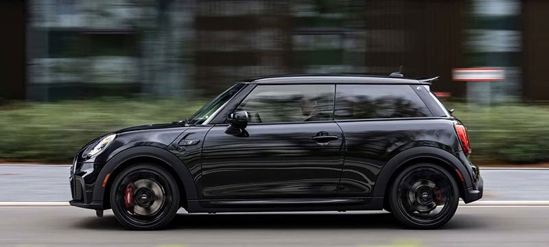 Side view of a MINI John Cooper Works 1 To 6 Edition vehicle driving alone on a street facing left, with bushes in the background along and the vehicle's surroundings blurred out.